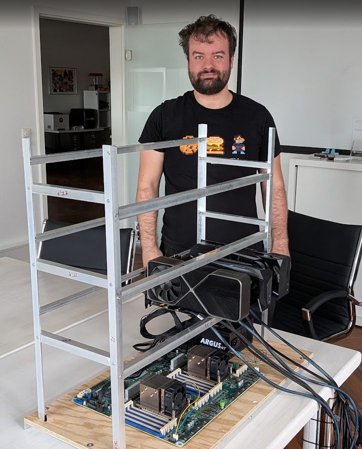 Aluminiumrahmen auf einer Holzplatte, darin das Dual-Sockel-Board mit zwei CPU-Kühlern; die ersten beiden Grafikkarten hängen bereits im Rahmen, dahinter steht ein Kollege.