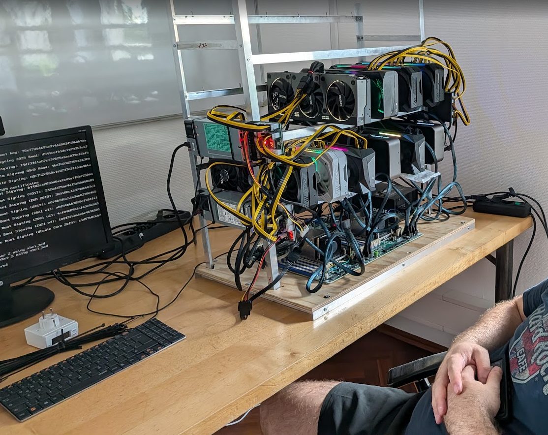 Das bestückte Rig auf einem Tisch: drei Ebenen voller Grafikkarten, gelbe Stromkabel, links die Netzteile, rechts daneben ein Monitor mit Bootmeldungen.