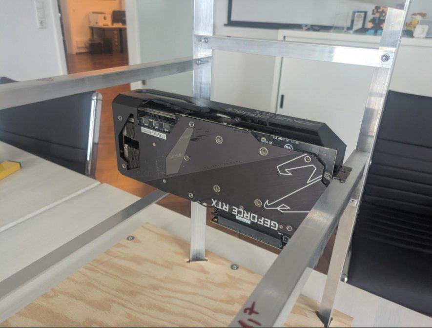Eine einzelne schwarze Grafikkarte hängt zur Probe in einem noch leeren Aluminiumrahmen, im Hintergrund ein Büroraum.