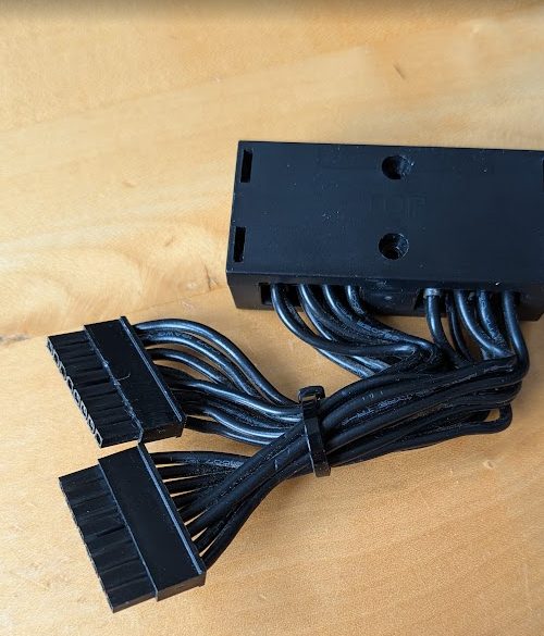 Die ausgebaute Strombuchsen-Baugruppe einer Grafikkarte auf einem Holztisch: ein schwarzer Kunststoffblock mit zwei PCIe-Stromsteckern und angeschlossenem Kabelbaum.